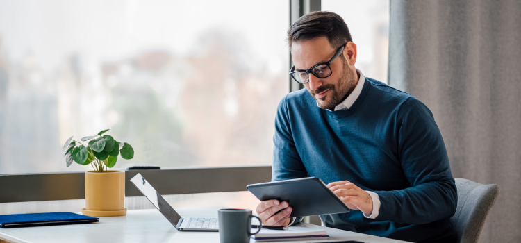 Image d'un homme assis à son bureau regardant sa tablette devant un ordinateur portable.